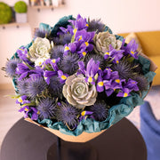 A close-up view of a stunning bouquet, showcasing the vibrant blue irises, purple thistle, and a succulent from above. The eco-friendly kraft paper peeks out subtly, drawing attention to the lush flowers and their beautiful arrangement.