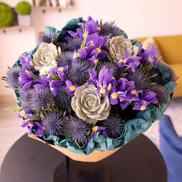 A close-up view of a stunning bouquet, showcasing the vibrant blue irises, purple thistle, and a succulent from above. The eco-friendly kraft paper peeks out subtly, drawing attention to the lush flowers and their beautiful arrangement.
