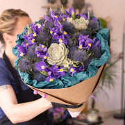 Stunning fresh flowers bouquet featuring vibrant blue irises, purple thistles, and a fresh succulent, elegantly wrapped in eco-friendly kraft paper with green tissue. A perfect blend of natural beauty and sustainability.