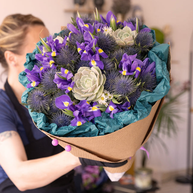 Stunning fresh flowers bouquet featuring vibrant blue irises, purple thistles, and a fresh succulent, elegantly wrapped in eco-friendly kraft paper with green tissue. A perfect blend of natural beauty and sustainability.