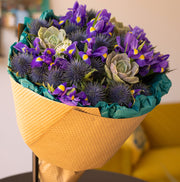 A beautifully wrapped bouquet featuring blue irises, purple thistle, and a succulent, elegantly arranged in eco-friendly kraft paper and green tissue. Captured from a different angle, the flowers' vibrant colors and textures stand out, creating a stunning visual that exudes natural charm and sustainable beauty.