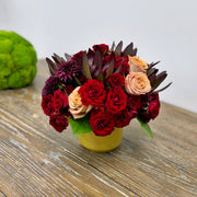 Side view of a fall bouquet in a Dijon-colored vase, featuring a lush arrangement of red spray roses, mums, cappuccino roses, and orange carnations, capturing the depth and texture of the flowers.