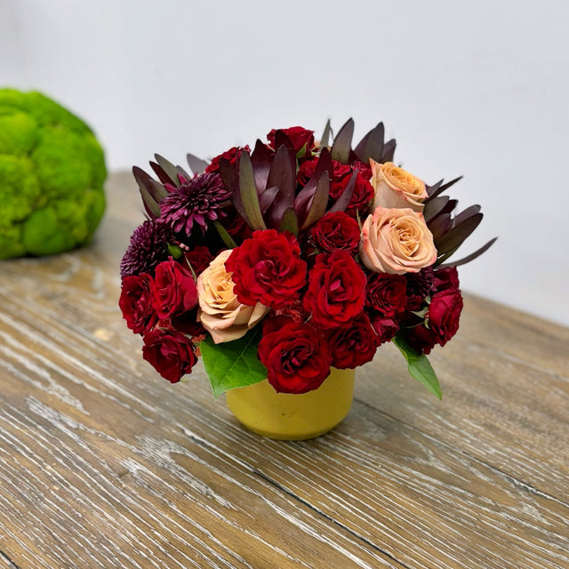 Side view of a fall bouquet in a Dijon-colored vase, featuring a lush arrangement of red spray roses, mums, cappuccino roses, and orange carnations, capturing the depth and texture of the flowers.