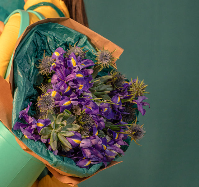 A stunning purple bouquet featuring vibrant blue irises, delicate purple thistle, and a striking succulent, wrapped in eco-friendly kraft paper and green tissue. A perfect blend of nature’s beauty and sustainable elegance.