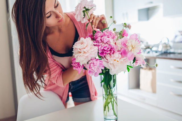 Woman puts peonies flowers in vase. Housewife taking care of coziness and decor on kitchen. Composing bouquet.