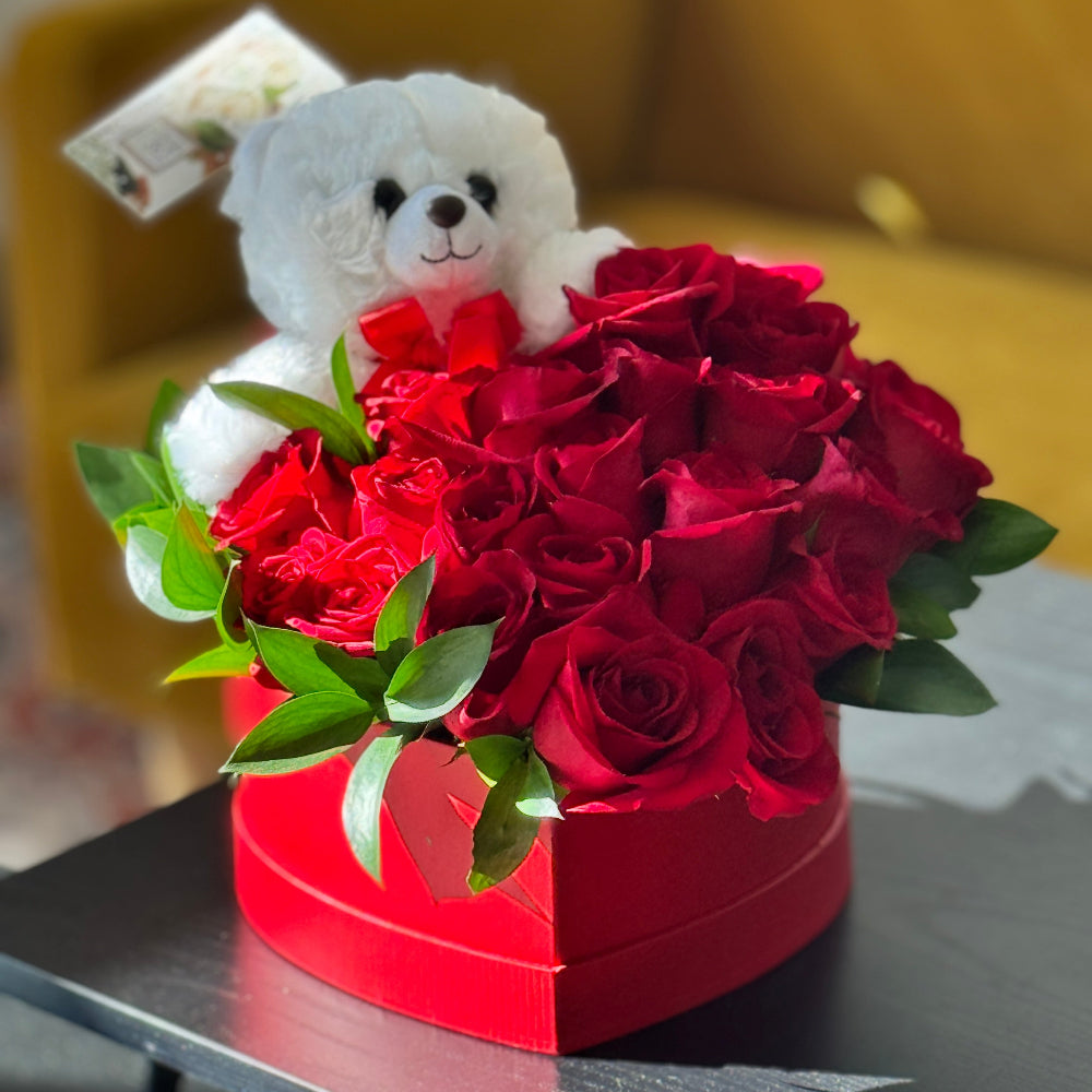White teddy bear sitting behind a heart-shaped box of red roses on a table.