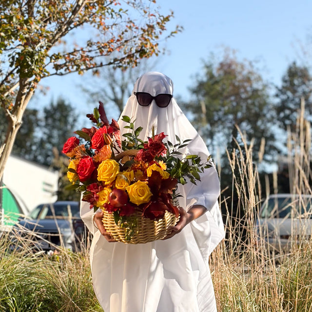A person wearing a white ghost costume and black sunglasses holds a large  basket arrangement with coral roses, yellow roses, decorative apples, and fall greenery