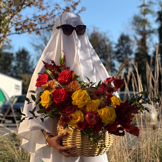 A person wearing a white ghost costume with sunglasses holds a large Fall basket centerpiece with yellow, red, and coral roses, chrysanthemums, and autumn foliage