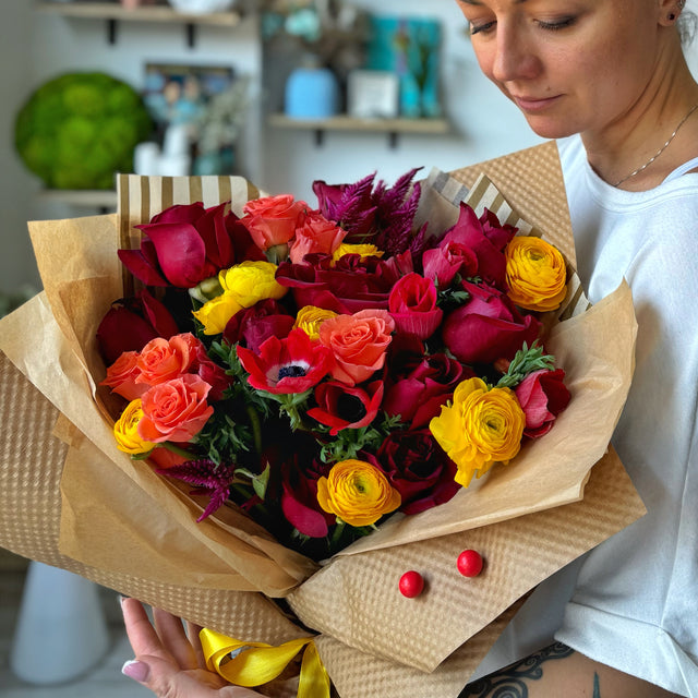 Florist hugging the Juicy Fall bouquet, which features vibrant yellow ranunculuses, red roses, orange spray roses, and red anemones, wrapped in kraft paper.