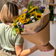 Florist presenting a cheerful bouquet of sunflowers and asters, elegantly wrapped in eco-friendly brown tissue and kraft paper.