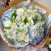 Close-up of a bouquet featuring blue hydrangeas, white roses, delicate baby's breath, and stock flowers, elegantly wrapped in craft paper.