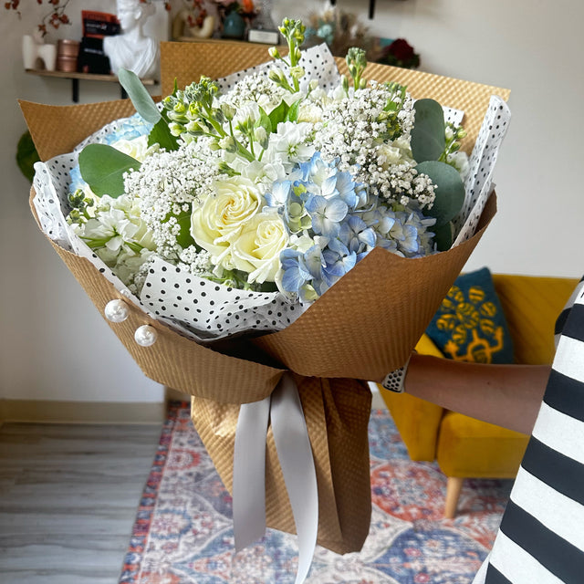 Florist holding a beautiful bouquet of blue hydrangeas, white roses, baby's breath, and stock flowers, showcasing its elegant arrangement.