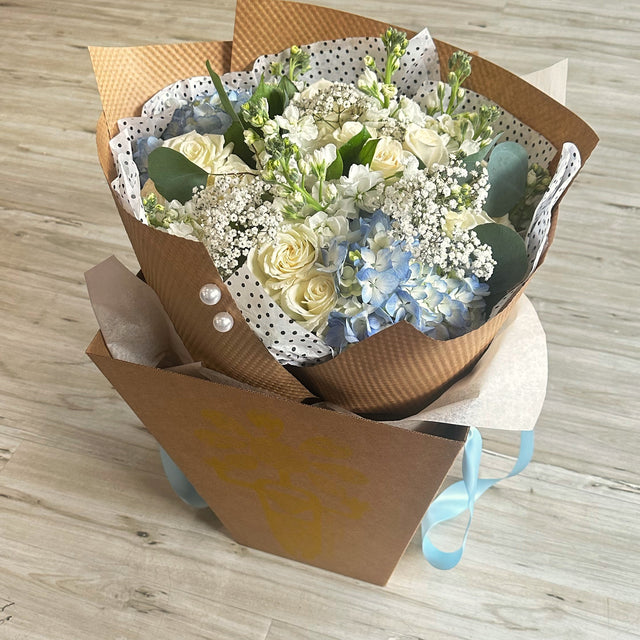 Elegant bouquet of blue hydrangeas, white roses, baby's breath, and stock flowers placed in a branded cone box for delivery.