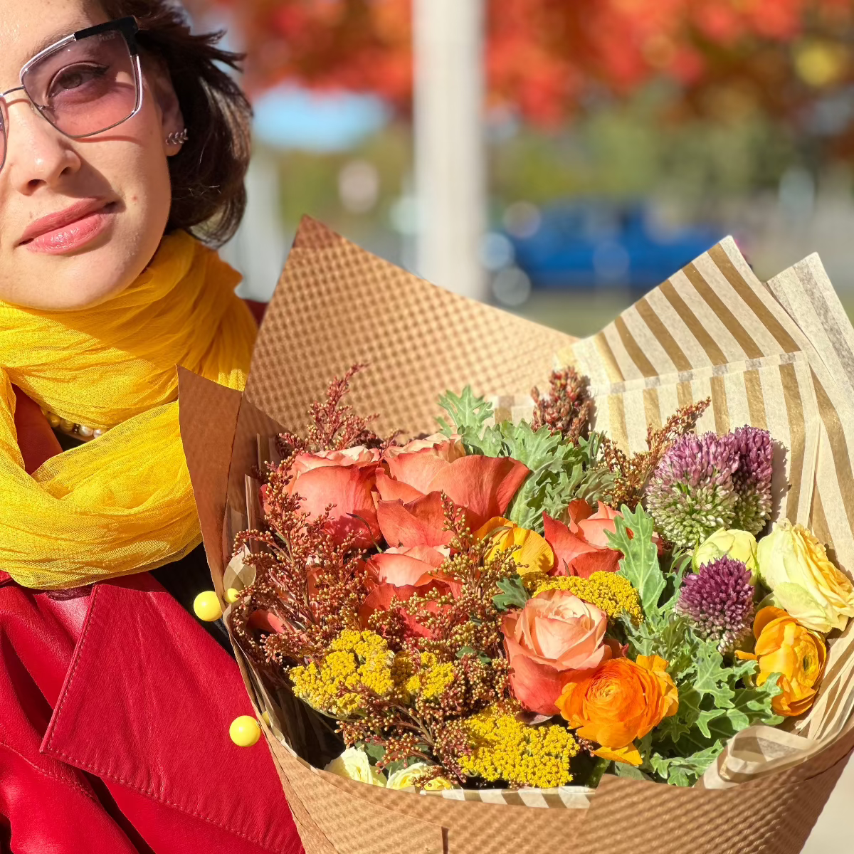 A close-up of a woman in bright clothing holding a vibrant fall-colored bouquet wrapped in rustic kraft paper.