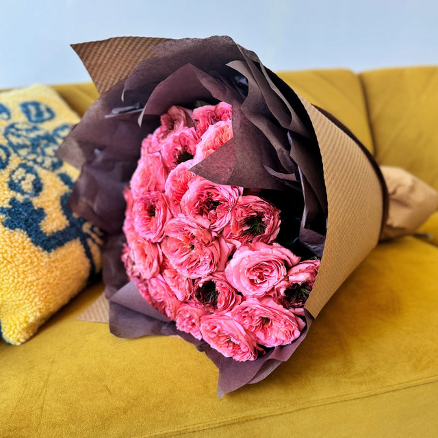 Bouquet of coral roses with large, fluffy heads and dark green centers, displayed on a couch, wrapped in brown tissue and kraft paper.