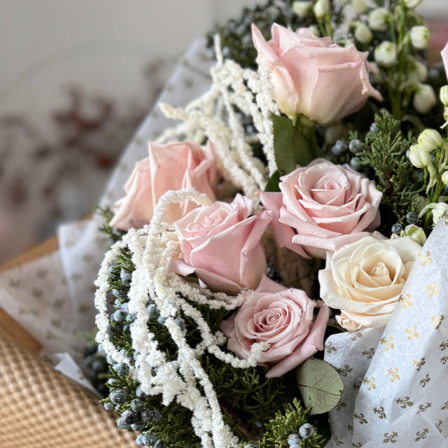 Bouquet of pink and white roses with greenery, wrapped in brown paper and lace, held by a person.