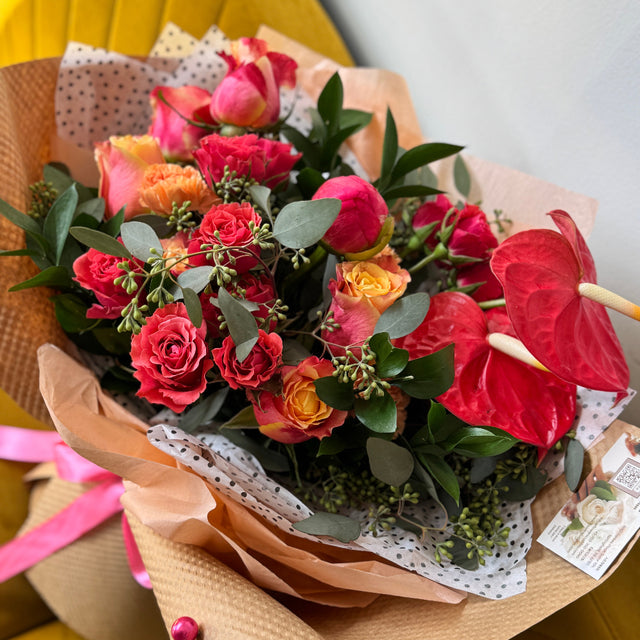 Bouquet of flowers with red and orange roses, anthuriums, and greenery on a yellow surface.