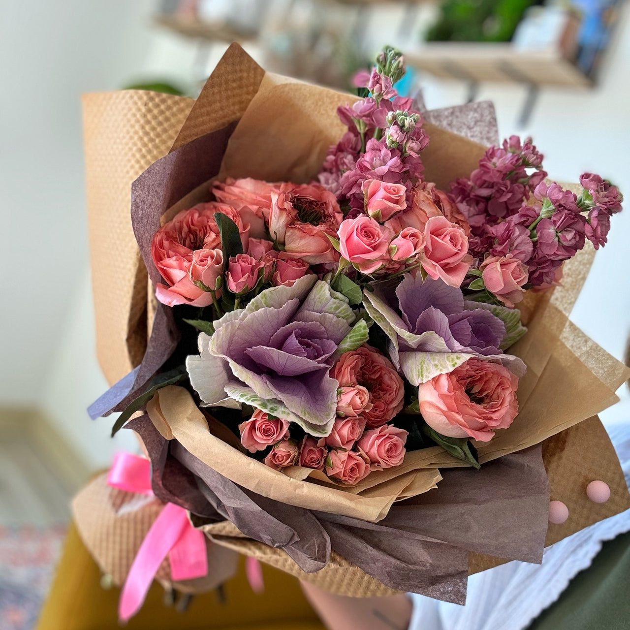 An open-faced Pink and purple fresh flower bouquet featuring Coral Expression roses, purple kale, pink spray roses, and mauve stock flowers, arranged in a one-sided design. The vibrant flowers are elegantly wrapped in kraft paper and craft tissue, with a pink detail ribbon adding a charming touch.