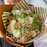 Close-up of the Champagne Dreams bouquet with cappuccino roses, white roses, and Queen Anne's lace, elegantly wrapped in striped brown and gold tissue paper and kraft paper, held by a florist.