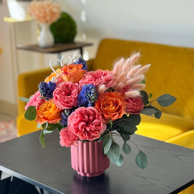 Colorful flower arrangement in a pink vase on a table with a yellow chair in the background.