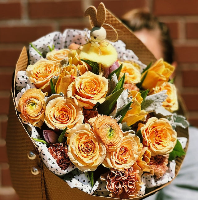 Yellow roses and ranunculuses wrapped in elegant cream tissue paper and kraft paper, with a small bunny toy nestled on top. The charming bouquet is beautifully presented, combining cheerful flowers with a cute decorative touch.