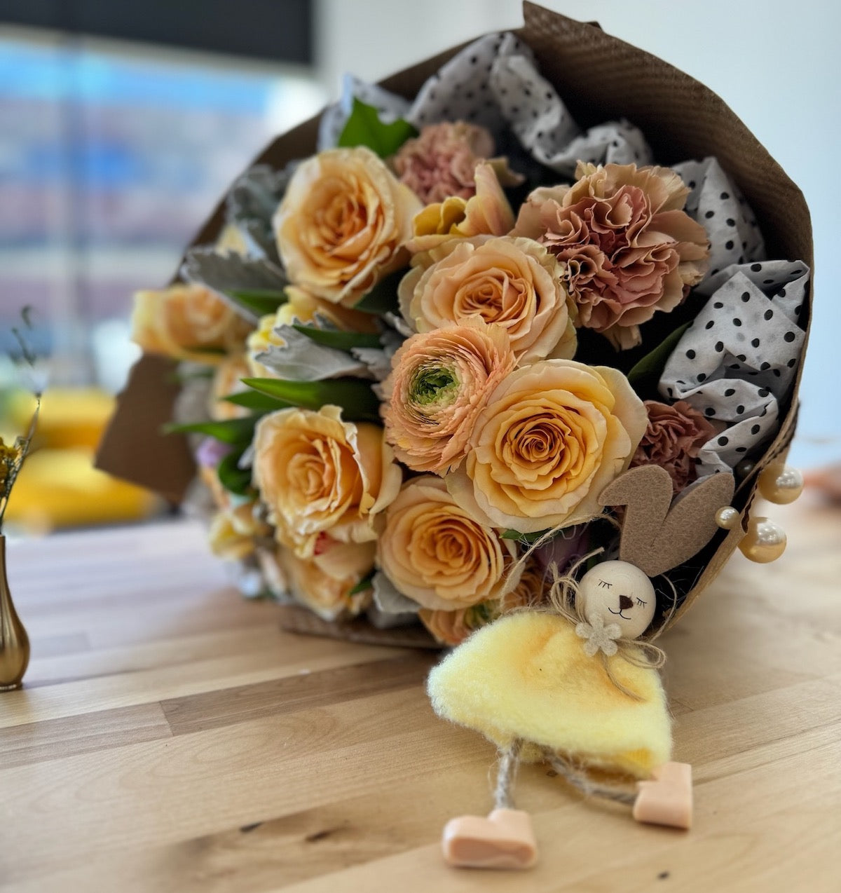 Flowers in yellow tones, including roses and ranunculuses, lying on a table with a small bunny toy leaning gently against the bouquet. The arrangement features warm, cheerful hues, creating a delightful and charming floral display.