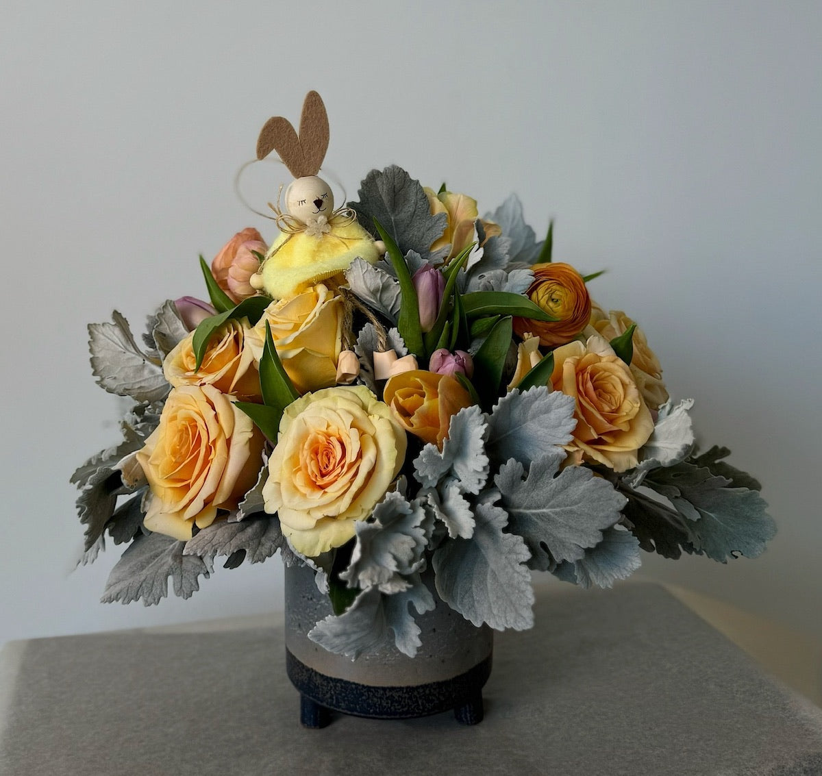 Yellow roses and ranunculuses arranged in a gray ceramic vase, with the bouquet topped by a small bunny toy. The arrangement also includes silver dusty miller grinnery, adding a touch of elegance and contrast to the cheerful floral display.