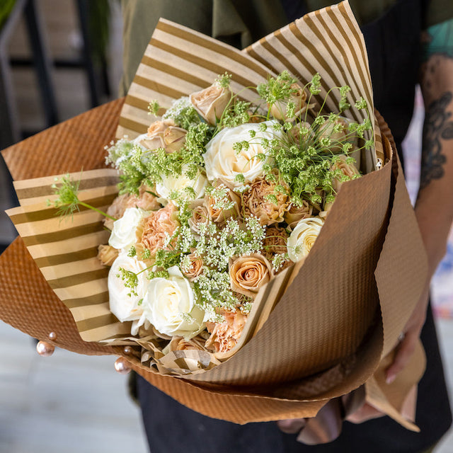 Close-up of the Champagne Dreams bouquet featuring cappuccino roses, white roses, and Queen Anne's lace, beautifully wrapped in brown and gold striped tissue and kraft paper, displayed in the hands of a florist.