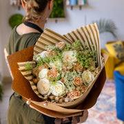 Champagne Dreams bouquet featuring cappuccino-colored roses, white roses, and Queen Anne's lace, beautifully wrapped in striped brown and gold tissue paper with kraft paper. The bouquet is delicately held by a florist, showcasing its elegant charm.