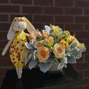 Yellow roses and ranunculuses arranged in a gray ceramic vase, with a large plush bunny named Dale standing next to the bouquet. The soft tones of the flowers and the plush bunny create a cozy and inviting display.