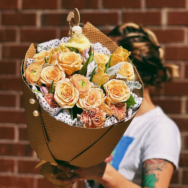 A person holding a wrapped bouquet of yellow roses and ranunculuses, wrapped in elegant cream tissue paper and kraft paper. The bouquet is beautifully presented, with the holder’s hands gently showcasing the vibrant flowers and charming presentation.