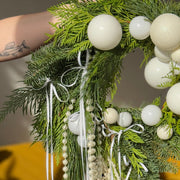Decorative wreath with greenery, white spherical ornaments, and ribbons on a blurred indoor background.