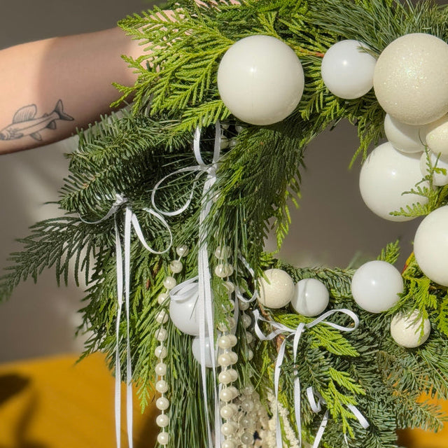 Decorative wreath with greenery, white spherical ornaments, and ribbons on a blurred indoor background.