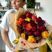 Juicy Fall bouquet held by a florist, featuring vibrant yellow ranunculuses, red roses, orange spray roses, and red anemones.