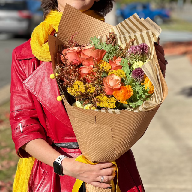 A woman in colorful attire holding a lush autumn bouquet wrapped in kraft paper, showcasing rich fall hues and textures.