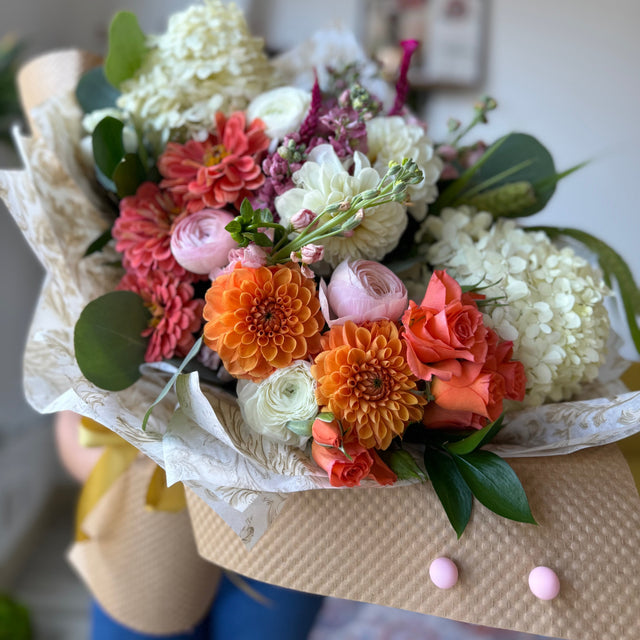 Close-up of the Seasonal Splendor Bouquet held by a florist, showcasing a mix of orange, pink, and cream white flowers.