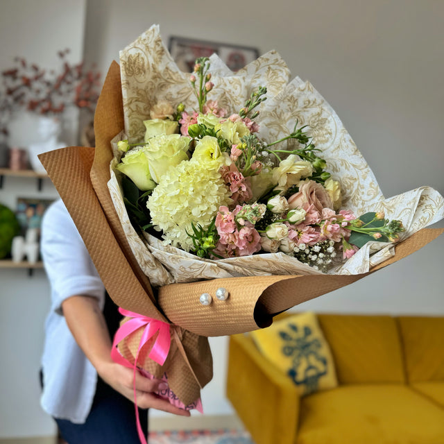 An elegant bouquet of white hydrangeas, white roses, ranunculus, and light pink stock flowers, fully visible in the florist's hands. The bouquet is wrapped in white tissue paper with a gold pattern.