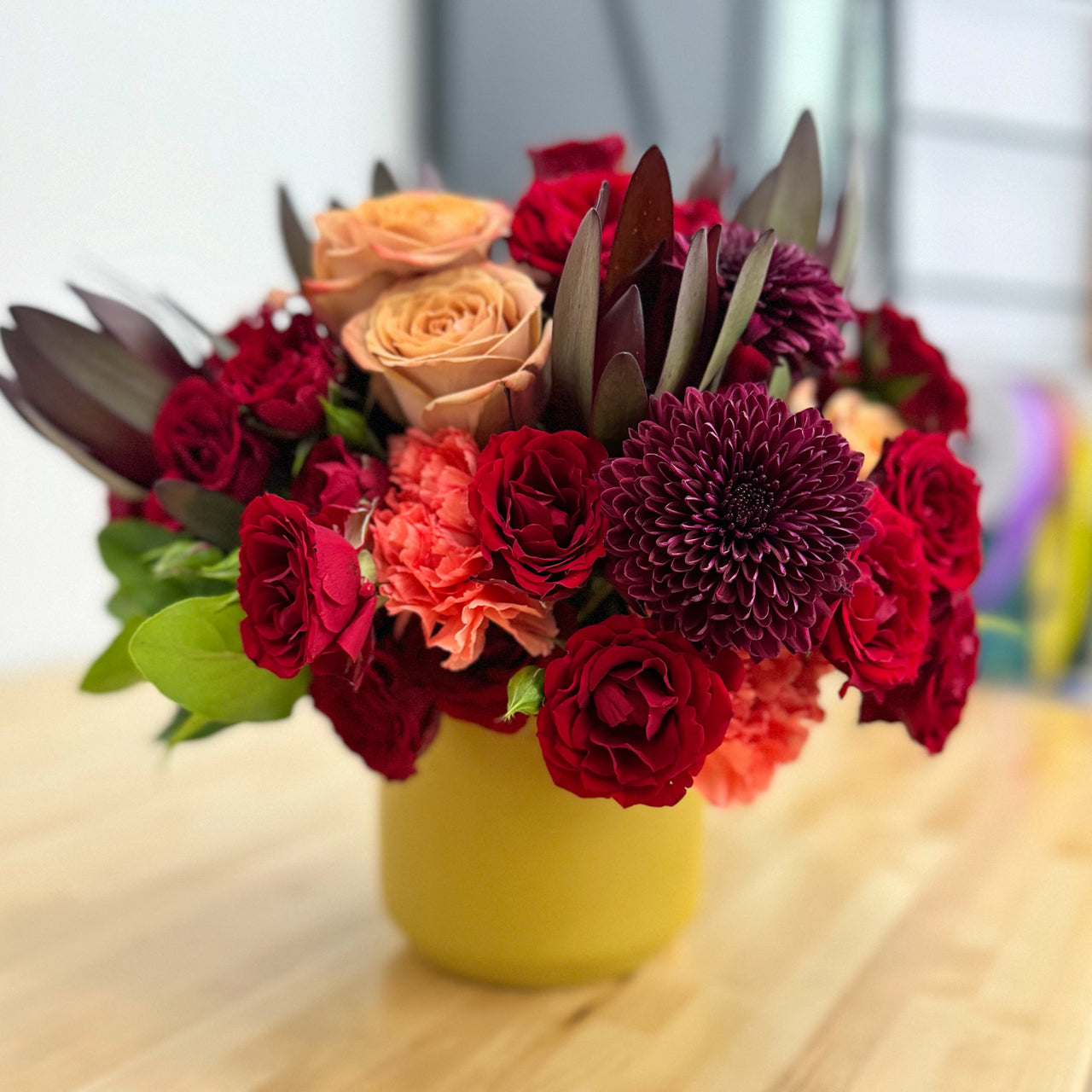 Fall bouquet in Dijon-colored vase featuring red spray roses, deep purple mums, cappuccino roses, and orange carnations.