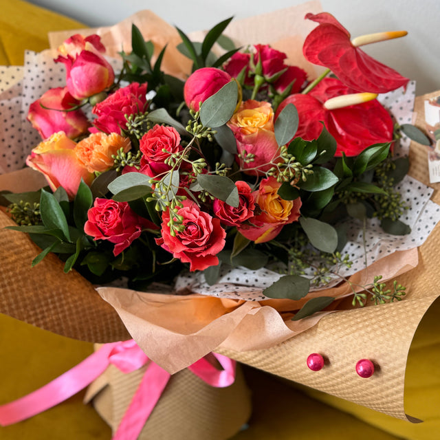 Bouquet of flowers with red roses, pink and orange flowers, and green leaves on a yellow surface.