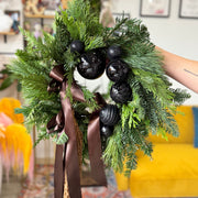 Green wreath with black ornaments and brown ribbons held by a person.