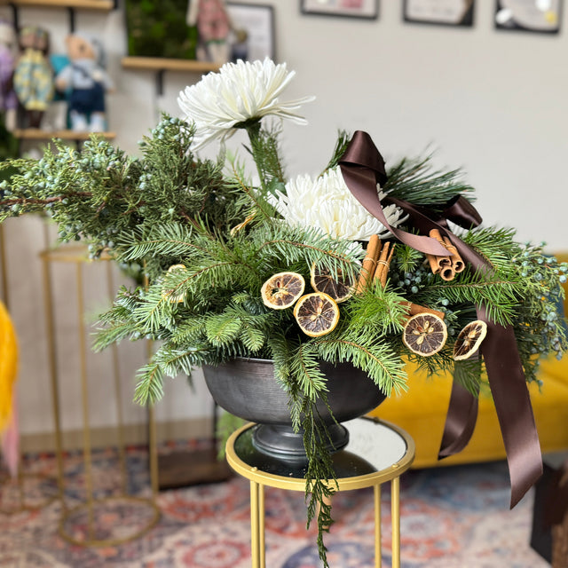Holiday floral arrangement with greenery, white flowers, and a brown ribbon in a living room setting.