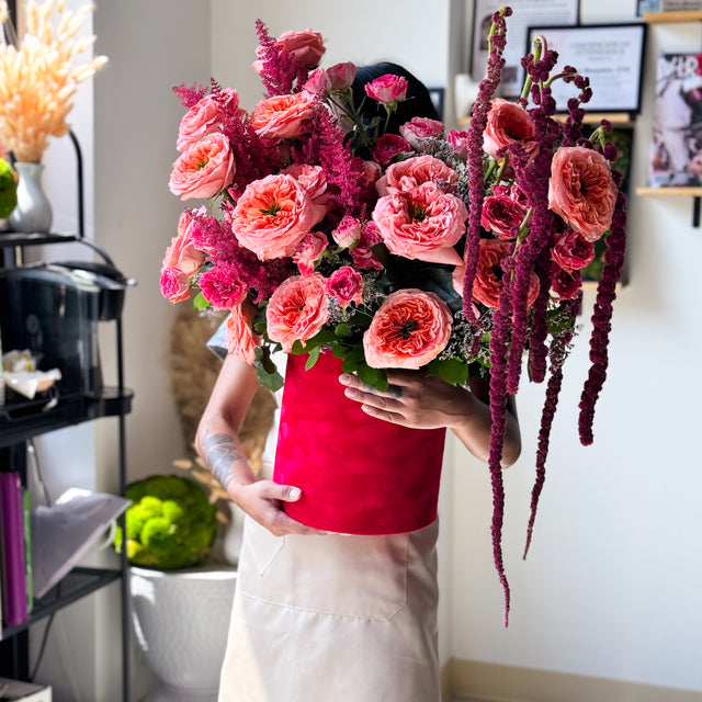 A person in a cream apron holds a large, vibrant floral arrangement in a bright red cylindrical container. The bouquet features pink and coral roses, deep magenta amaryllis, and green foliage, set against a softly blurred indoor background.