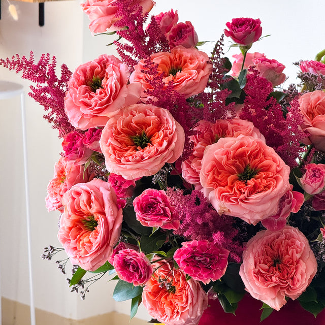 A vibrant floral arrangement featuring large ruffled pink and coral roses, deep magenta astilbe, and green foliage, presented in a bright indoor setting.