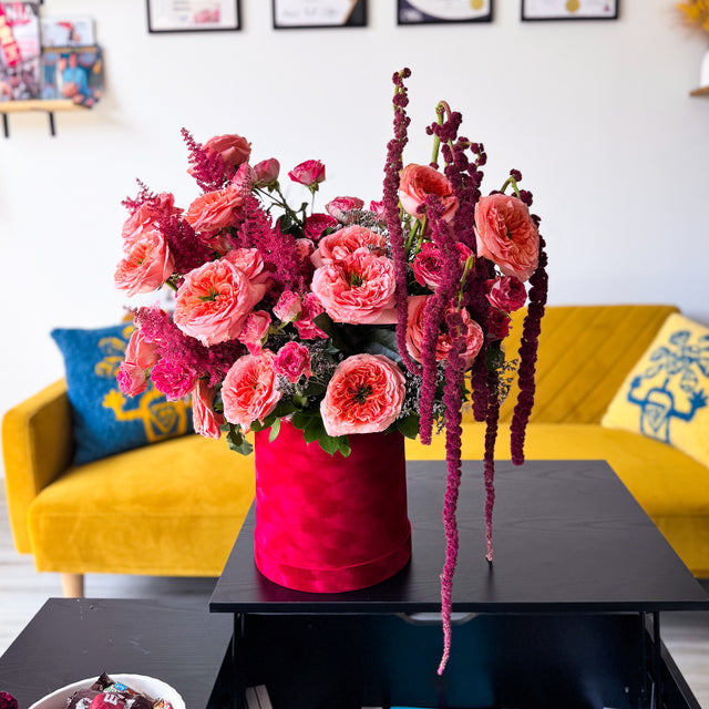 A vibrant floral arrangement featuring pink and coral ranunculus, deep magenta amaryllis, and burgundy spikes, presented in a textured red cylindrical pot on a black coffee table in a bright, modern living room.