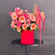 A vibrant floral arrangement featuring pink and peach roses, pink astilbe, and long hanging burgundy amaryllis, presented in a textured bright red cylindrical pot on a round black table against a dark gray background.