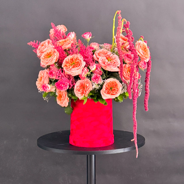 A vibrant floral arrangement featuring pink and peach roses, pink astilbe, and long hanging burgundy amaryllis, presented in a textured bright red cylindrical pot on a round black table against a dark gray background.