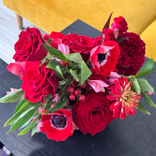 A vibrant red floral arrangement featuring red roses, anemones, and pink flowers with green leaves, displayed on a dark table in a modern living room setting with a yellow sofa in the background.