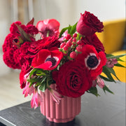 A vibrant red floral arrangement featuring roses, anemones, and carnations in a ribbed pink ceramic vase, displayed on a dark wooden table in a softly blurred indoor setting.