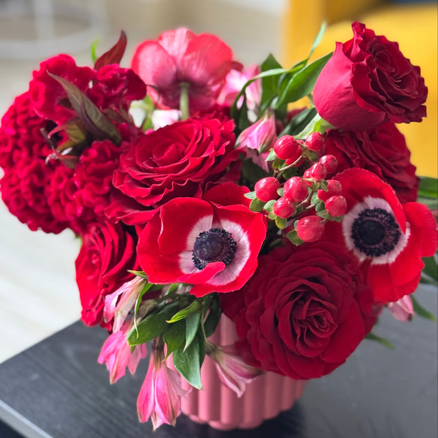 A vibrant red flower bouquet featuring roses, anemones, and carnations arranged in a pink ribbed vase, resting on a dark table with a softly blurred indoor background.