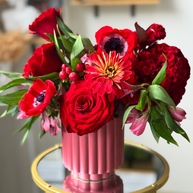 A vibrant floral arrangement featuring red roses, red anemones, and a yellow-and-red zinnia in a pink ribbed ceramic vase, displayed on a gold-rimmed round table in a softly blurred indoor setting.